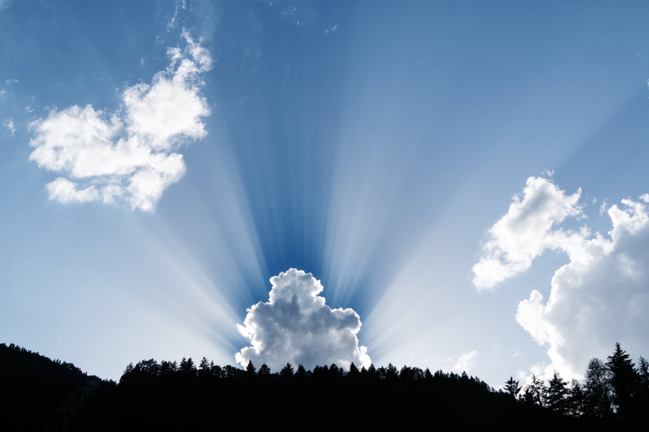 A stunning natural phenomenon as sunrays pierce through fluffy clouds above a silhouetted forest.
